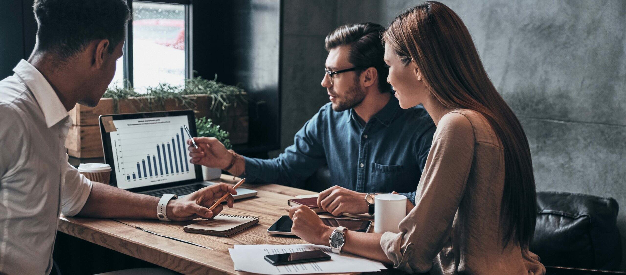 Três pessoas (um homem branco, um homem negro e uma mulher branca) sentadas em volta de uma mesa de escritório de madeira, observando um grafico que representa o crescimento das franquias de edcucação para 2023
